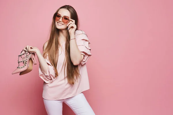 Menina modelo loira bonita e feliz com um sorriso brilhante em blusa bege e em óculos de sol rosa na moda segurando sapatos elegantes em suas mãos e posando no fundo rosa, isolado — Fotografia de Stock