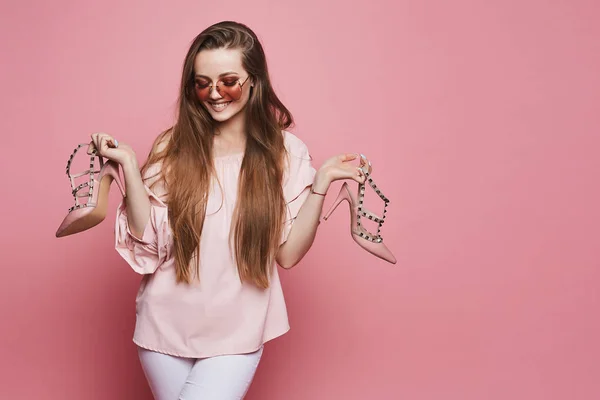 Felice ragazza modello bionda con un sorriso lucido in camicetta beige e in occhiali da sole rosa alla moda tenendo scarpe alla moda nelle sue mani e posa sullo sfondo rosa, isolato — Foto Stock