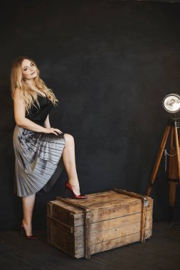 Siyah Saten bluz ve koyu kırmızı ayakkabılar, gümüş etek büyük meme ile güzel ve şık sarışın model kız, ahşap bir göğüs üzerinde duruyor, gülümseyen ve koyu iç stüdyoda poz