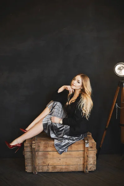 Beautiful and fashionable blonde model girl in silver skirt, in black jacket and dark-red shoes, sits on a wooden chest, smiling and posing in studio at dark interior