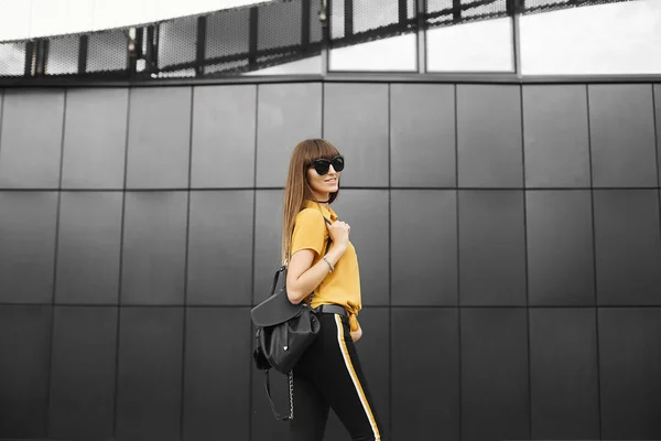 Menina modelo de cabelos castanhos bonita na camisa sem mangas amarela e nos óculos de sol da moda com mochila na moda sorrisos e posando ao ar livre no fundo urbano — Fotografia de Stock