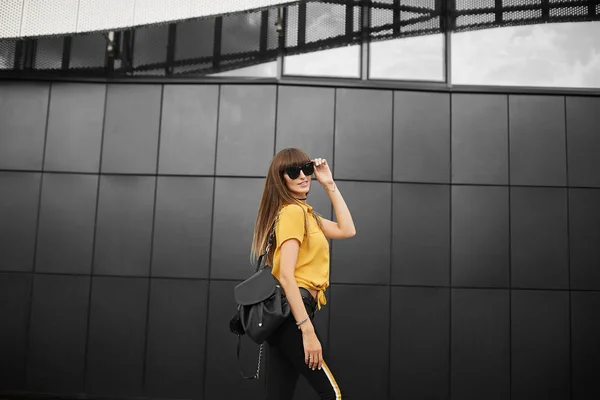 美丽的棕色头发模特女孩在黄色无袖衬衫和时尚太阳镜与时髦的背包微笑, 并在城市背景摆出户外 — 图库照片