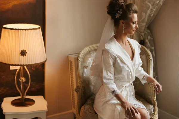 Beyaz giyinme elbisesi içinde parlak makyaj ile güzel esmer model kız sandalyeye oturur ve iç poz, Genç gelin düğün hazırlanması — Stok fotoğraf