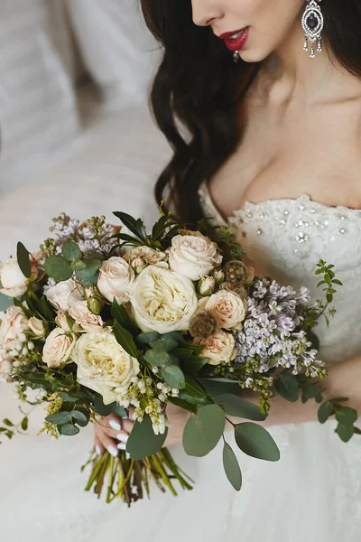 A big bouquet of flowers in the hands of the beautiful busty brunette model girl with big luxury earrings with diamonds in the fashionable wedding dress