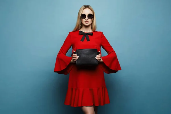 Divatos és szép modell szőke lány, professzionális smink, stílusos napszemüveg és elbűvölő piros ruhában, fekete íj és fekete tengelykapcsoló táska a kezében pózol kék háttér — Stock Fotó