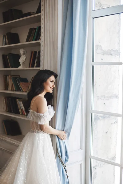 Sinnlich und verführerisch brünette Modell Mädchen in stilvollem Spitzenkleid mit nackten Schultern blickt durch das Fenster und posiert in der Nähe Bücherregal auf Luxus-Interieur — Stockfoto