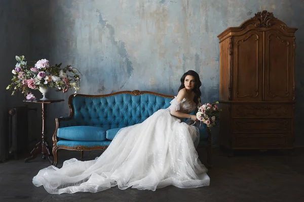 Sexy et belle fille modèle brune dans une robe de mariée en dentelle élégante et à la mode avec des épaules nues se trouve sur le canapé antique et posant dans un intérieur vintage de luxe — Photo