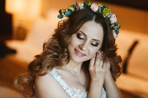 Mooie brunette model meisje met heldere make-up en met stijlvolle bruiloft kapsel versierd met bloemen legt op haar oorbellen en poseren op interieur — Stockfoto