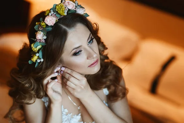 Beautiful brunette model girl with bright makeup and with stylish wedding hairstyle decorated with flowers puts on her earrings and posing at interior — Stock Photo, Image