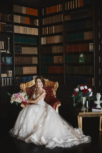 Mulher jovem morena elegante com maquiagem brilhante e com brincos elegantes em vestido de noiva na moda segurando o buquê de flores e posando no interior vintage de luxo — Fotografia de Stock