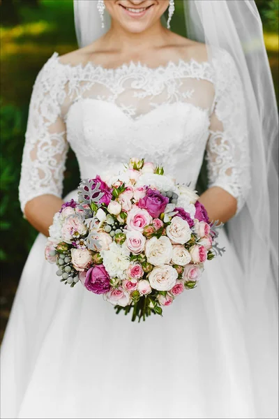 Fashionable model girl with a shiny smile and with stylish earrings in trendy wedding dress holding the bouquet of colorful flowers — Stock Photo, Image