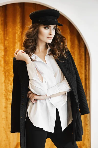 Fashionable and beautiful brunette model girl with gentle makeup in the trendy hat, in stylish oversize jacket and in a white unbuttoned blouse posing at interior Stock Image