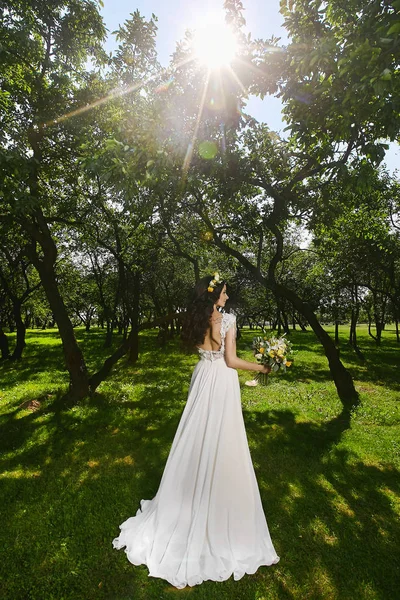 Sposa felice con una corona floreale in testa e in un abito da sposa con schiena nuda che tiene un mazzo di fiori nel giardino verde nella giornata di sole — Foto Stock