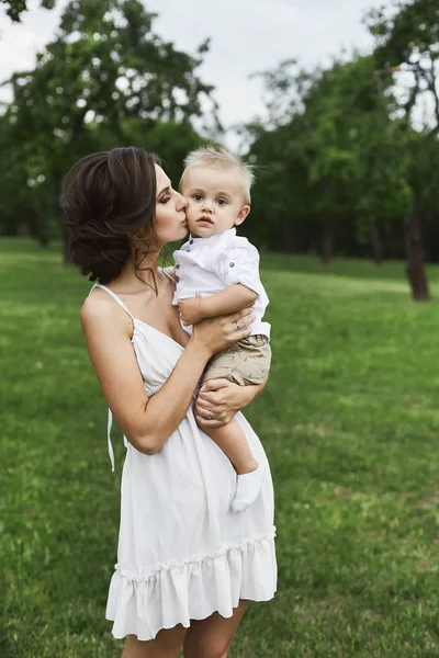 Hermosa morena joven mamá en vestido blanco corto elegante sosteniendo en sus manos y besando a su lindo bebé al aire libre —  Fotos de Stock