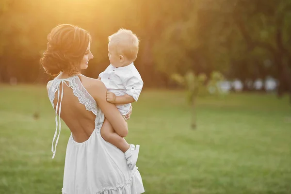 Schöne brünette model girl, junge mama in kurzen weißen stilvollen kleid hält auf ihren händen ihr süßes happy baby boy und posiert im grünen park bei untergang — Stockfoto