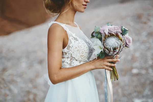 Menina modelo loira bonita com penteado de casamento modelagem em um vestido de renda branca na moda com um buquê de flores exóticas em suas mãos — Fotografia de Stock