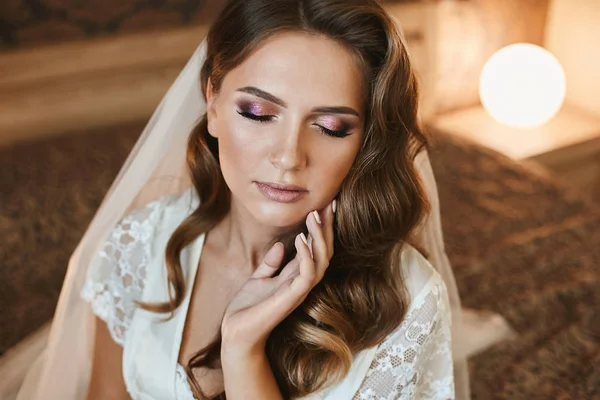 Mulher de cabelos castanhos bonita com penteado de casamento e maquiagem brilhante senta-se com os olhos fechados na cama — Fotografia de Stock