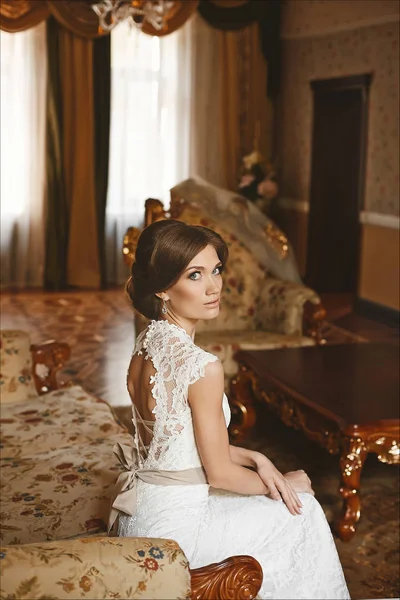 Elegáns fiatal menyasszony, gyönyörű kék szemek és az esküvői frizura a stílusos ruha csipke ül a szüreti kanapé luxus belső — Stock Fotó
