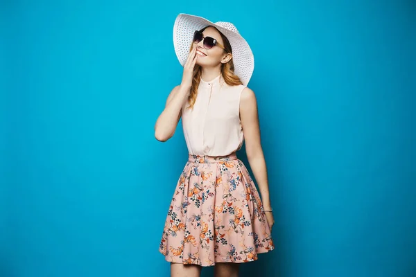 Красива і модна блондинка модель дівчина в бежевій блузці, рожева спідниця, стильні сонцезахисні окуляри і капелюх посміхається і позує в студії на синьому фоні — стокове фото