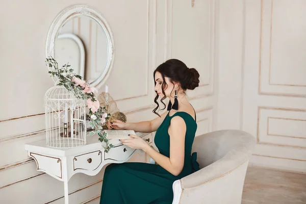 Mulher morena elegante com penteado elegante em vestido de noite elegante e em sapatos da moda senta-se à mesa de vestir — Fotografia de Stock