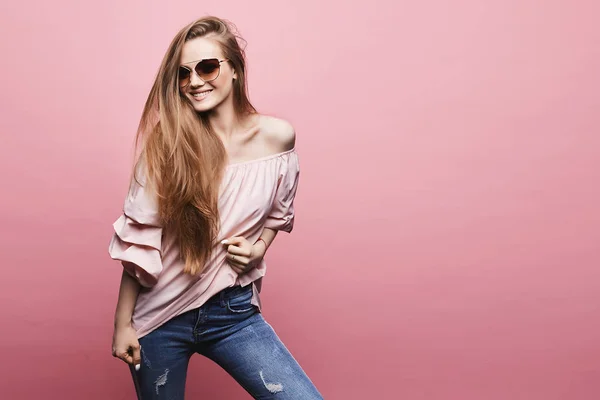 Happy and emotional young woman with blond long hair and with perfect body in stylish sunglasses and in the fashionable outfit at the pink background, isolated — Stock Photo, Image
