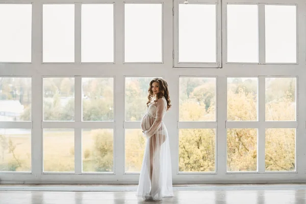 Gyönyörű fiatal terhes nő divatos frizura egy stílusos peignoir mosolygós és pózol a nagy ablak közelében a fény belső — Stock Fotó