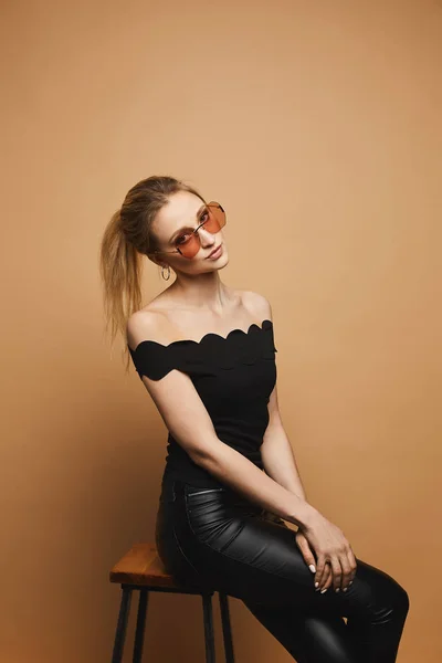 Mulher bonita nova com corpo magro perfeito em calças de couro preto, em uma blusa preta e em óculos de sol na moda no fundo laranja, isolado — Fotografia de Stock
