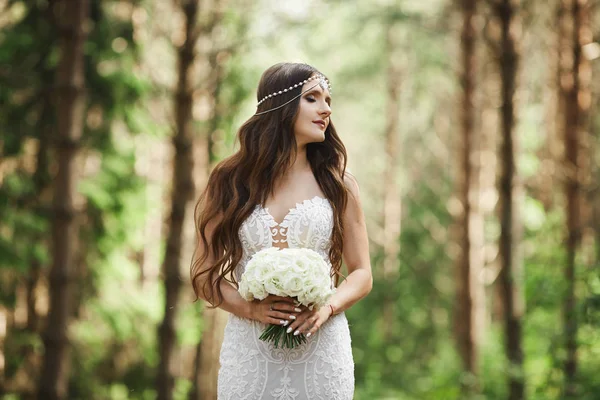 Élégante femme modèle brune avec coiffure de mariage avec bijoux en robe de dentelle tenant un bouquet de fleurs dans ses mains et posant à la forêt tôt le matin — Photo