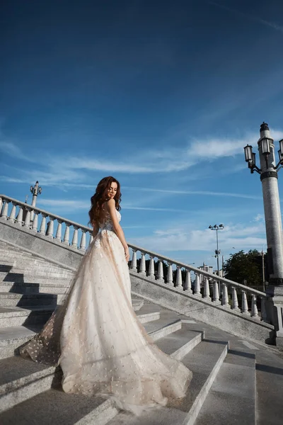 Mooie model vrouw met perfect lichaam in luxe trouwjurk staat met terug op de trap en poseren met blauwe hemel op de achtergrond — Stockfoto