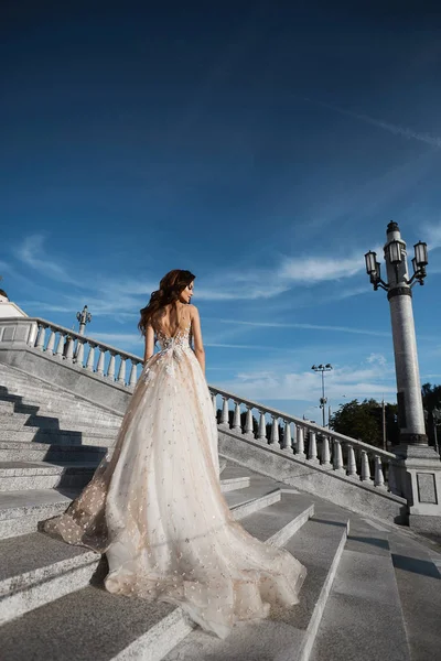 Schöne Modell Frau mit perfektem Körper in Luxus-Hochzeitskleid steht mit dem Rücken auf der Treppe und posiert mit blauem Himmel im Hintergrund — Stockfoto