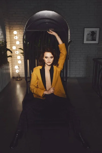 Sexy jovem mulher no blazer amarelo no corpo nu e na moda calças quadriculadas senta-se em uma cadeira de metal e posando no interior escuro — Fotografia de Stock