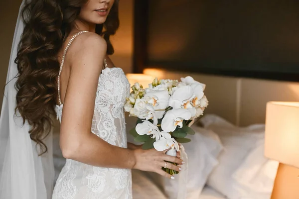 Mulher morena nova com penteado de casamento em um vestido de noiva de renda com um buquê de flores frescas em suas mãos em interior elegante — Fotografia de Stock