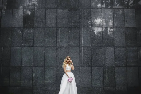 Junge und schöne blonde Frau mit Blumenstrauß in der Hand in einer stylischen Bluse und im Tüllrock, die draußen vor der schwarzen Granitwand posiert — Stockfoto