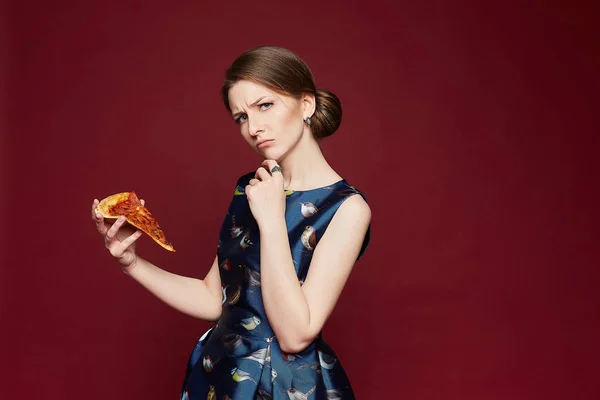 Vacker brunett modell kvinna med blå ögon i en fashionabel blå klänning håller en bit pizza i handen och tänker om att äta denna pjäs, isolerad vid röd bakgrund — Stockfoto