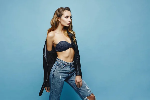 Mulher modelo jovem bonita com lábios cheios sexy e com corpo perfeito em uma jaqueta de couro e em jeans na moda posando no fundo azul, isolado — Fotografia de Stock