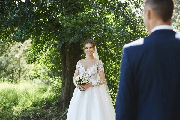 Hermosa joven con una bonita sonrisa en el elegante vestido de novia sosteniendo un ramo de flores en las manos y mirando a su novio al aire libre —  Fotos de Stock