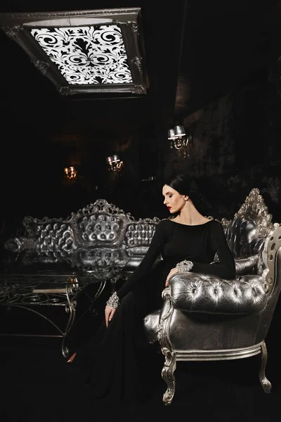 Moda e sexy donna modello bruna con corpo perfetto e con le labbra rosse in abito da sera nero con si siede sul divano d'argento al moderno interno di lusso — Foto Stock