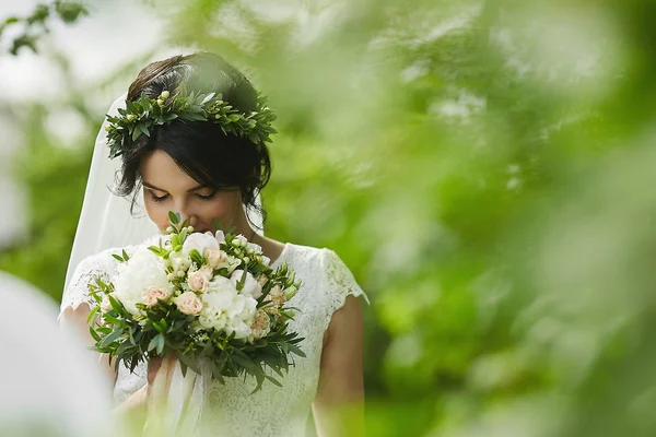 Young beautiful bride with green floral wreath in her wedding hairstyle enjoys a bouquet of rose flowers outdoors in summer day