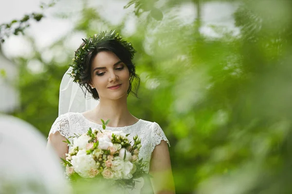Young beautiful bride with green floral wreath in her wedding hairstyle enjoys a bouquet of rose flowers outdoors in summer day