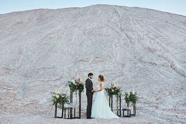 Ein schönes Liebespaar in einer weißen Wüste, eine junge Frau mit Hochzeitsfrisur in einem stilvollen Kleid und ein gutaussehender Mann im grauen Anzug, der bei der Hochzeitszeremonie posiert — Stockfoto