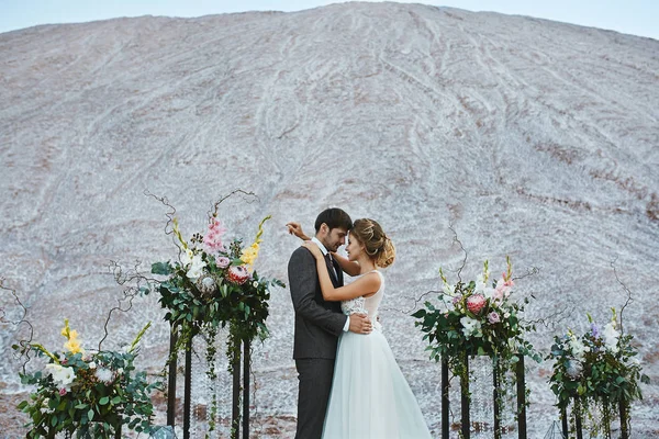 Ein schönes Liebespaar in einer weißen Wüste, eine junge Frau mit Hochzeitsfrisur in einem stilvollen Kleid und ein gutaussehender Mann im grauen Anzug, der bei der Hochzeitszeremonie posiert — Stockfoto