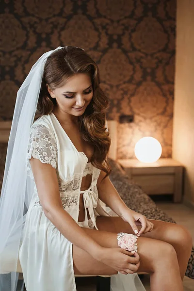 Sexy donna modello bruna mezza nuda con acconciatura da sposa e con corpo perfetto in lingerie di pizzo elegante si mette la giarrettiera nuziale e si prepara per una cerimonia di nozze — Foto Stock