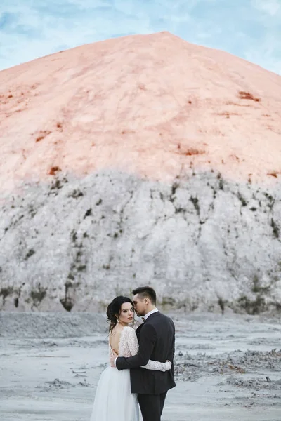 A beautiful couple of lovers at the white salt desert, a young model woman in the wedding dress and handsome brutal man in the suit with the white mountain at the background — Stock Photo, Image