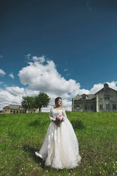 Schöne Modell Frau in stilvollem Hochzeitskleid hält einen Blumenstrauß und posiert auf dem grünen Gras mit einem alten Gebäude im Hintergrund — Stockfoto