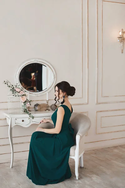 Giovane donna alla moda modello con acconciatura elegante in abito da sera alla moda si siede sulla poltrona vicino alla toletta — Foto Stock