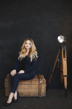 Koyu mavi takımlı mavi gözlü genç sarışın model iç kısımdaki ahşap sandıkta oturuyor.