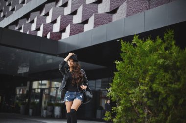 Güzel ve moda esmer model kadın çorap, kot şort ve deri ceket, siyah sırt çantası onu şık şapka ayarlama ve yaz şehir Caddesi tarafından yürüyüş