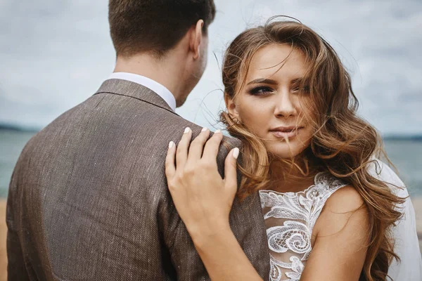 Coppia alla moda e bella di abbracci appena sposati al molo sulla costa, bella giovane donna con trucco luminoso e con i capelli selvaggi abbraccia un uomo bello alla moda — Foto Stock