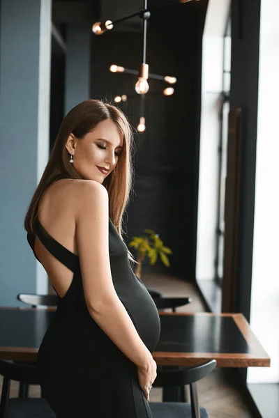 Mulher grávida bonita e jovem em um vestido de noite preto elegante posando no interior escuro minimalista do café de luxo — Fotografia de Stock