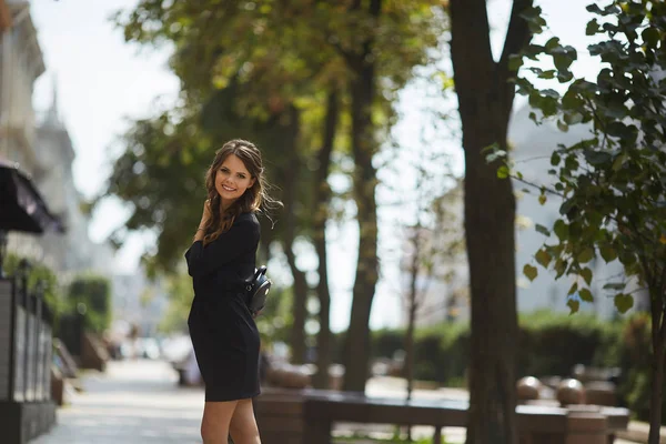 Krásná a módní modelová dívka s lesklým úsměvem ve stylových černých krátkých šatech, které se za slunečného dne kráčejí v letní městské ulici — Stock fotografie